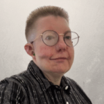 White person with a skin fade style hair cut wearing glasses, a hearing aid with green earmold and a black button up shirt with white vertical stripes facing to the right but with head turned forwards towards the camera